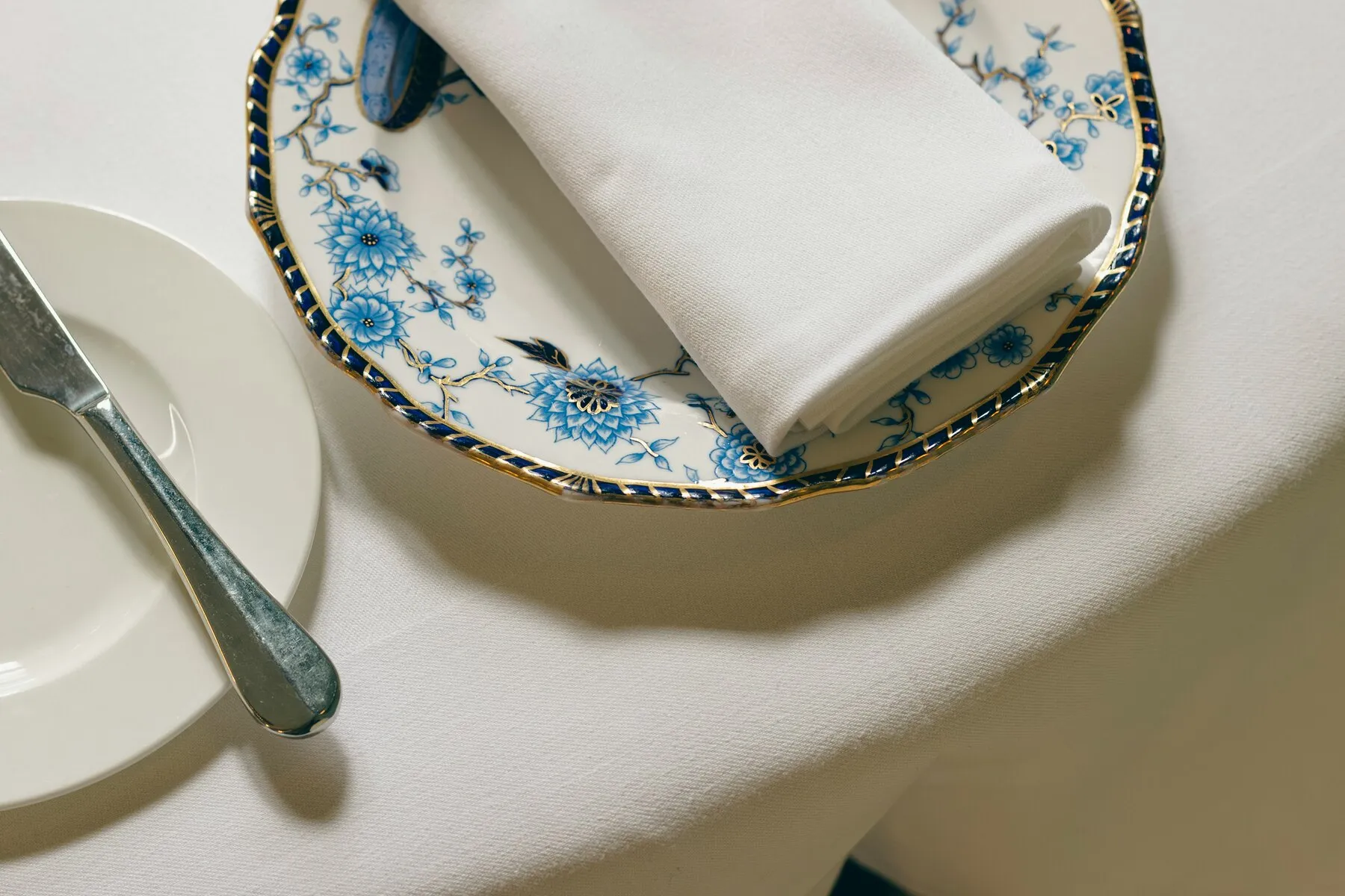 Gedekte tafel met strak geplooid wit servet op een bord met blauw-goud dessin, op wit tafellinnen